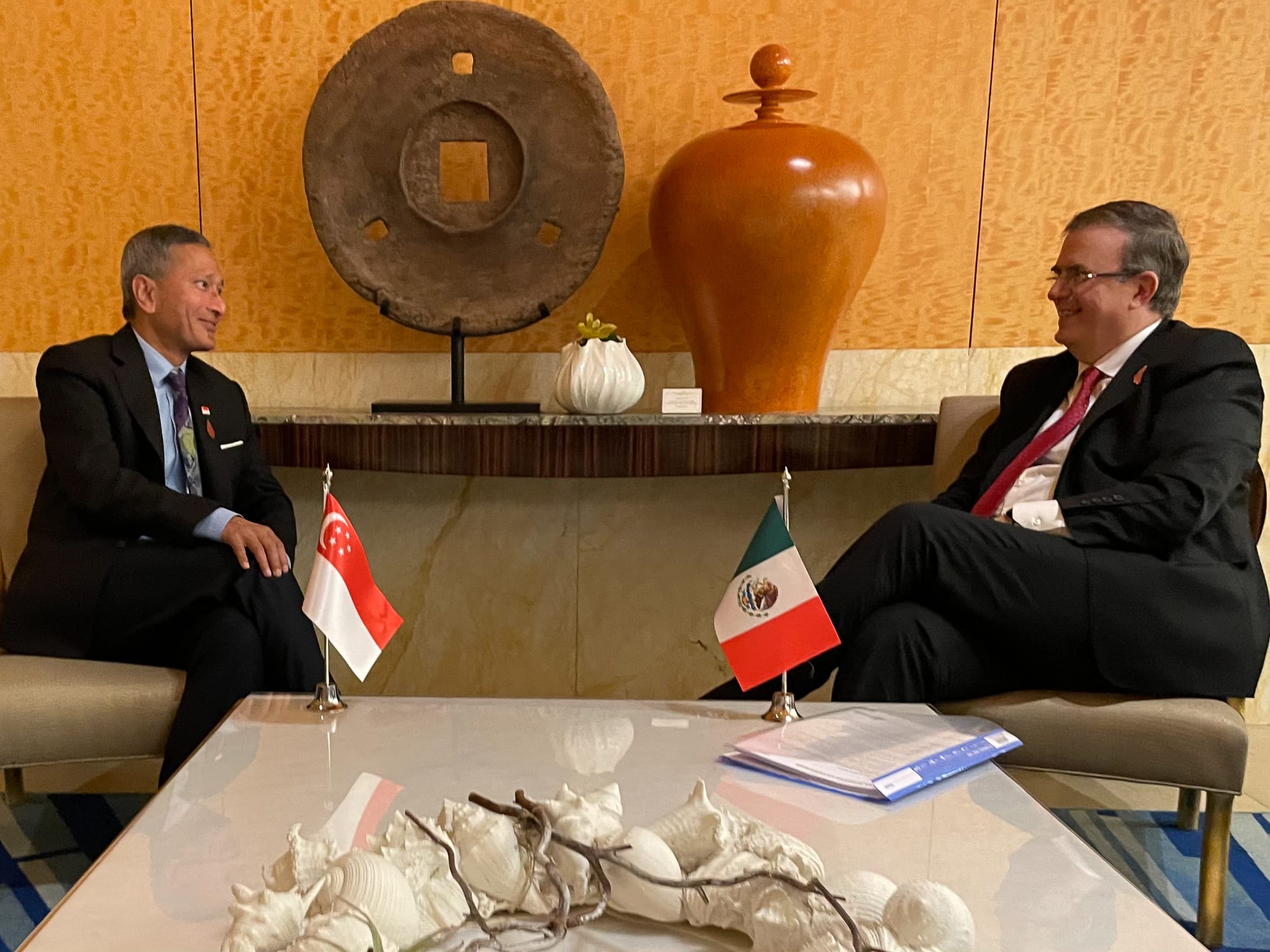 Two men in suits seated with Singapore and Mexico flags on a table with shells.